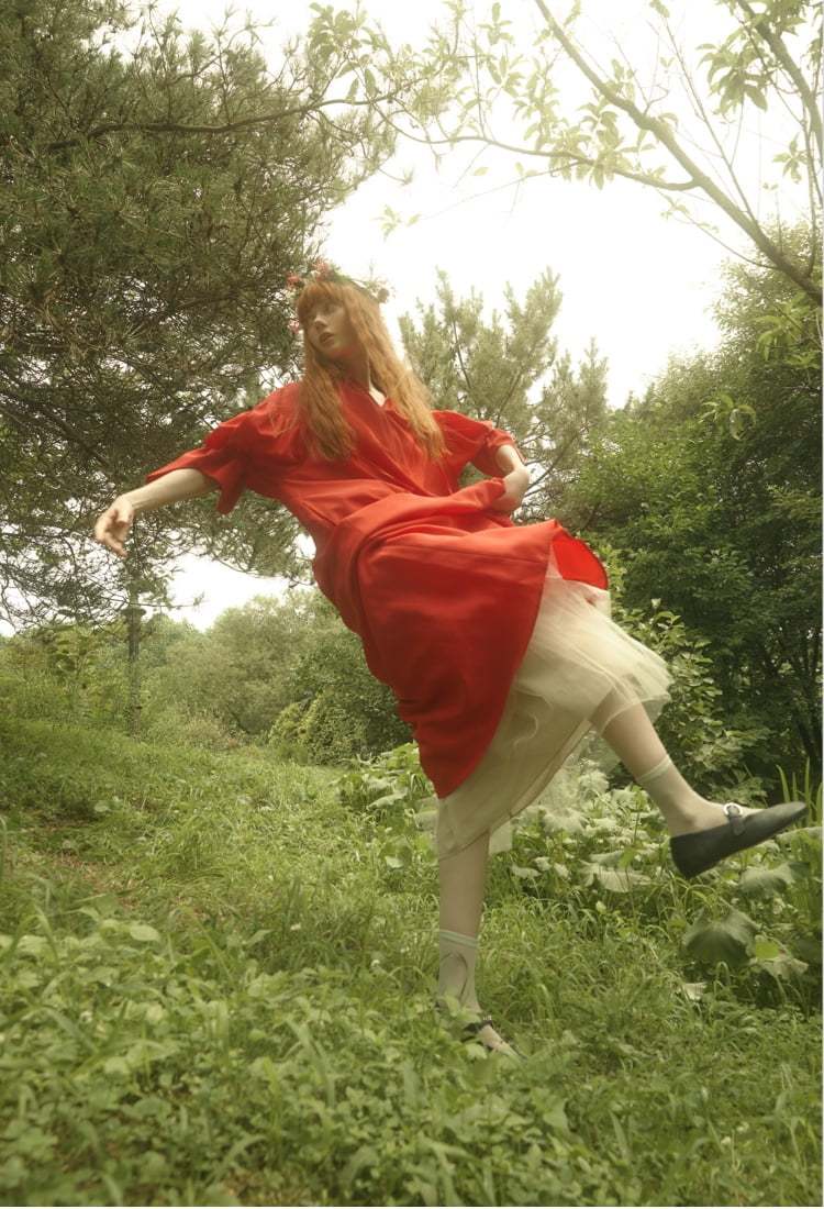 하얀 속치마를 입고 그 위에 엔폴드의 빨간 원피스 그리고 리포메이션의 검정 발레 슈즈를 신고 붉은 꽃 화관을 쓴 채 숲속에서 포즈를 취하고 있는 붉은 머리 외국 여성 모델