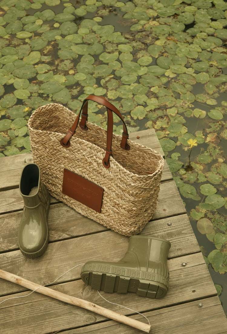 연못 위 나무판자에 놓여진 엠포리오 아르마니의 밀짚 소개의 브라운 가죽끈이 달린 가방과 어그의 녹색 앵클 레인부츠
